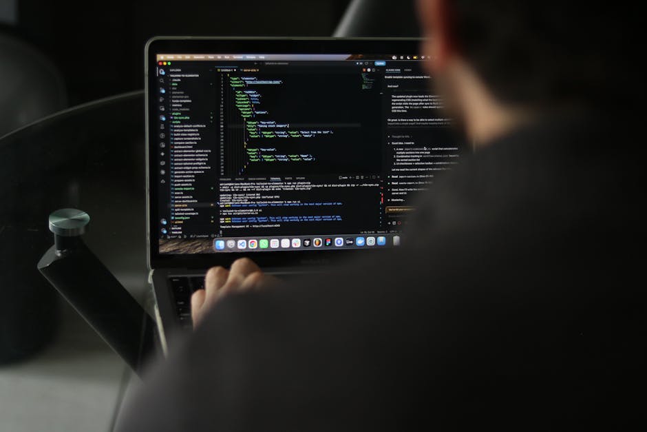 A person sitting in a vehicle, viewed from behind, working on a laptop with a dark screen displaying lines of color-coded code. The individual is partially visible, with blurred facial features, and i