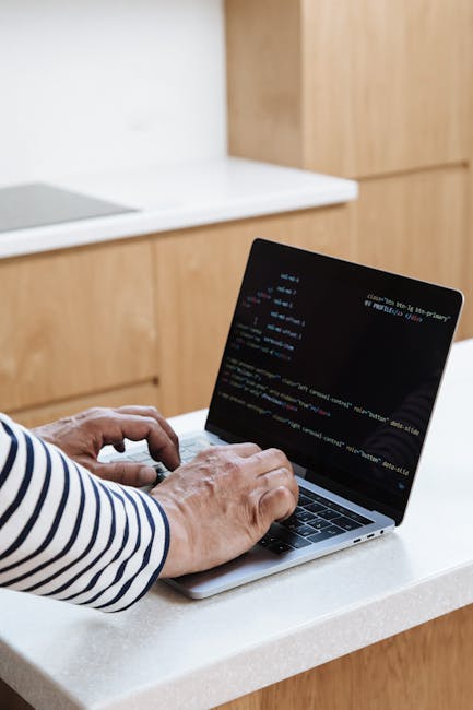 A person sitting in a vehicle, viewed from behind, working on a laptop with a dark screen displaying lines of color-coded code. The individual is partially visible, with blurred facial features, and i
