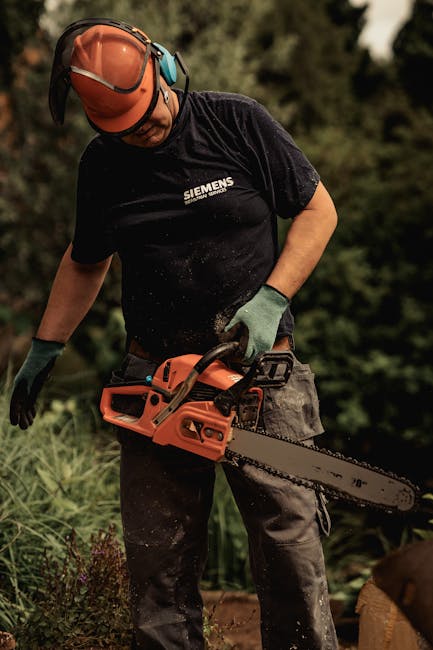 A gardener wearing a black Siemens T-shirt, protective orange helmet with a face shield, and green gloves is outdoors operating an orange chainsaw to trim or cut wooden or shrubbery material. The back